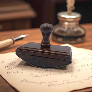 May include: A dark wooden ink blotter with a rounded handle sits on a piece of aged paper with handwritten script. An inkwell and pen are in the background, suggesting a vintage writing setup. The blotter is rectangular with a dark, polished finish.