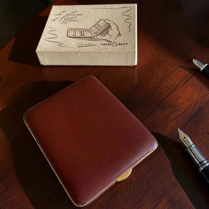 May include: A rectangular, dark brown, textured case with a gold-toned metal edge, accompanied by a beige box featuring the text "Le plaisir d'Offrir..." and "TREMBLAY" with a line drawing of a hand holding a similar case, plus a fountain pen.