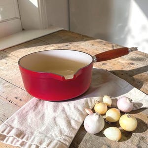 May include: A bright red enameled saucepan with a white interior, a pouring spout, and a long wooden handle. The pan is positioned on a light-colored fabric cloth next to a small cluster of whole yellow and purple onions on a wooden surface.