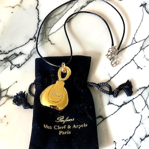 May include: A gold-colored pendant necklace with a black cord, featuring a pendant with the word "First" engraved on it. The necklace is displayed with a black velvet drawstring bag with the words "Parfums Van Cleef & Arpels Paris".
