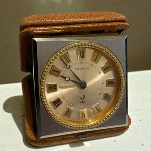 May include: A vintage travel clock with a square face and a gold-tone bezel. The clock face is a light gold color with black Roman numerals and hands. The clock is encased in a brown leather case. The text "DISCRETO" is visible on the clock face.