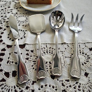 Puede incluir: Cuatro utensilios de servir plateados: un cuchillo de mantequilla, una espátula, una cuchara ranurada y un tenedor de servir. Cada utensilio tiene un diseño de concha en la base del mango. Una rebanada de pastel está sobre un plato blanco con borde dorado.