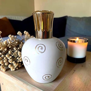 Könnte beinhalten: Eine weiße Duftlampe aus Keramik mit goldenen Spiralmustern und einem goldfarbenen Metallaufsatz. Eine brennende Kerze in einem Glas und Trockenblumen befinden sich ebenfalls auf dem Holztisch. Die Lampe ist ein dekorativer Artikel.