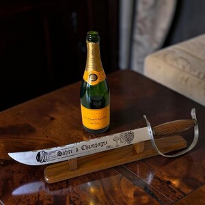 May include: A champagne bottle with a gold label and a green glass body sits on a wooden table next to a champagne saber. The saber has a wooden handle and a silver blade with the words "Sabre a Champagne" engraved on it.