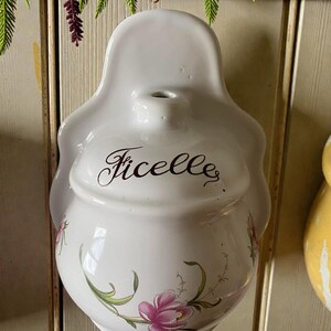 May include: White ceramic wall-mounted container with the word "Ficelle" in script. The container has a bulbous base with pink floral designs and green leaves. The top has a small opening.