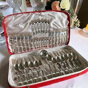 May include: A silver-toned flatware set housed in a red velvet-lined case with white satin fabric. The collection includes various sizes of forks, spoons, and knives, all featuring intricate embossed patterns on the handles, arranged neatly in rows.