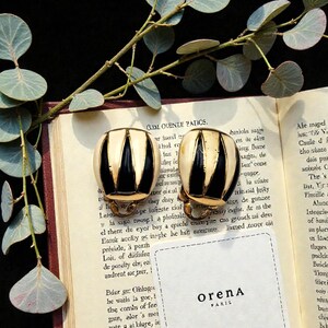 Peut inclure: Paire de boucles d'oreilles vintage à clip, avec un motif rayé noir et crème. Les boucles d'oreilles sont posées sur un livre ouvert, avec une branche de verdure et une petite carte blanche avec le texte "OrenA PARIS".