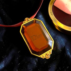 May include: A gold-framed pendant necklace with a large, amber-colored rectangular stone. The word "Dior" is subtly engraved on the stone. The necklace has a brown ribbon and is set against a dark background. Additional small amber-colored stones adorn the frame.