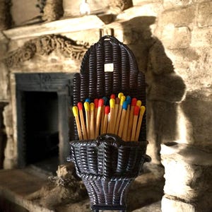May include: A dark brown wicker match holder filled with matches. The matches have red, yellow, and blue tips. The holder is shaped like a basket and is sitting on a stone surface.