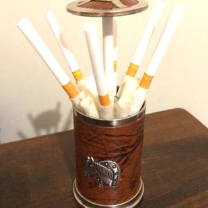 May include: A vintage cigarette holder with a brown leather-effect exterior and silver-coloured trim. It has a metal horse emblem and a decorative lid. Cigarettes with orange filters are visible inside the holder.