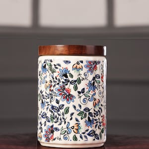 Può includere: Un barattolo cilindrico in ceramica con coperchio in legno. Il barattolo è decorato con un motivo floreale blu, rosso e arancione su sfondo crema. Il coperchio in legno è di colore marrone scuro.