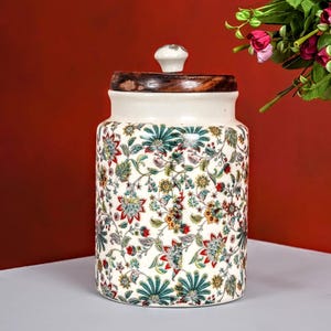 Op de afbeelding: Een keramische pot met een houten deksel. De pot is wit met een bloemmotief in groen, rood en geel. De deksel is donkerbruin met een kleine witte knop. De pot wordt waarschijnlijk gebruikt voor opslag.