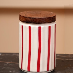 Può includere: Un barattolo di ceramica bianca con strisce rosse verticali e un coperchio rotondo in legno scuro. Il barattolo ha una forma cilindrica ed è adatto per la conservazione in cucina. Il design è semplice e moderno.
