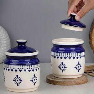 Puede incluir: Dos tarros de cerámica con tapas, con base color crema y detalles azul marino. Los tarros están decorados con un patrón de corazones y diseños florales. Se levanta una tapa, revelando el interior.