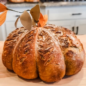 May include: A golden brown pumpkin-shaped loaf of bread with leaf-shaped designs on the top. The bread is sitting on a wooden cutting board.