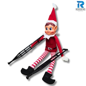 May include: A Christmas elf doll in a red outfit, white collar, and striped legs. The elf wears a red hat and black shoes. The elf is holding two black crutches. The Rodgers Design & Print logo is in the upper right corner.