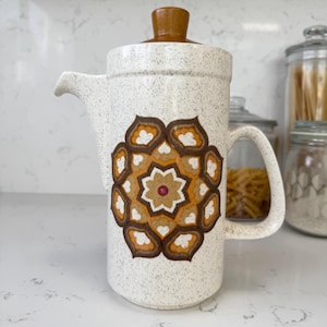 May include: A white ceramic coffee pot with a brown lid and a brown, orange, and white geometric design. The pot has a handle and a spout.