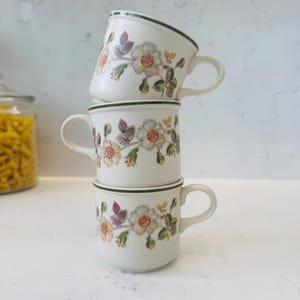 May include: A stack of three white ceramic mugs featuring a delicate floral pattern with yellow, pink, and green leaves. Each mug has a dark green rim and a small handle. The mugs are arranged vertically, with the top mug tilted slightly to the side.