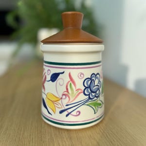 May include: A white ceramic jar with a floral design in shades of blue, yellow, pink, and green. The jar has a wooden lid.
