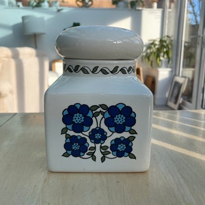 May include: White ceramic jar with a blue and green floral design. The jar has a white lid with a rounded top.