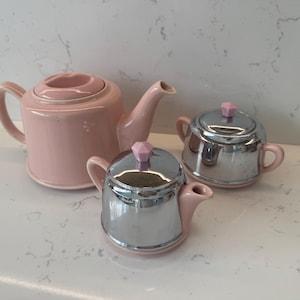 May include: A tea set consisting of a light pink ceramic teapot, a smaller light pink and chrome creamer, and a light pink and chrome sugar bowl. Each piece features a matching light pink, diamond-shaped knob on the lid.