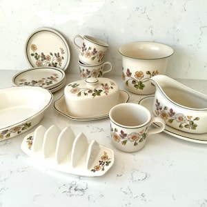 May include: A set of white ceramic dishes with a floral pattern in shades of green, brown, and pink. The set includes a butter dish, a gravy boat, a serving dish, a cup and saucer, and a toast rack.
