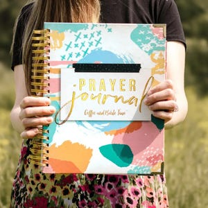 May include: A spiral-bound prayer journal featuring a colorful abstract cover with teal, orange, and pink brushstrokes. The center white label displays the gold foil text PRAYER journal and Coffee and Bible Time, accented by a black dotted strip.