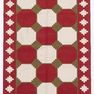 May include: A rectangular woven rug featuring a repeating geometric pattern of red and off-white octagons set against an olive green background. The rug is framed by a red border with white cross motifs and finished with off-white fringe at both ends.