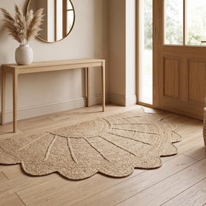 May include: A semi-circular rug made of woven natural tan fibres, featuring a sunburst pattern with radiating lines and a scalloped edge. The rug is laid flat on a light-coloured wooden floor, showcasing a textured, braided construction throughout.