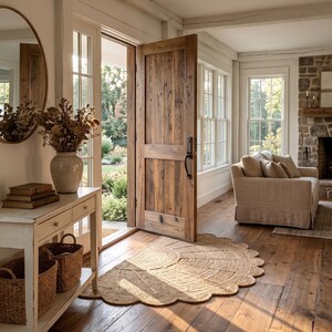May include: A semi-circular jute rug with a scalloped edge sits on a wooden floor in an entryway. The space features a white console table with two woven baskets, a vase of dried flowers, a round mirror, and a large wooden door left open.