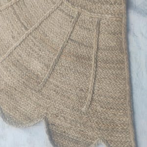May include: A tan woven jute rug featuring a scalloped edge and a geometric pattern created by raised braided lines. The natural fibre material has a textured appearance with a light brown colour palette throughout the entire surface area.