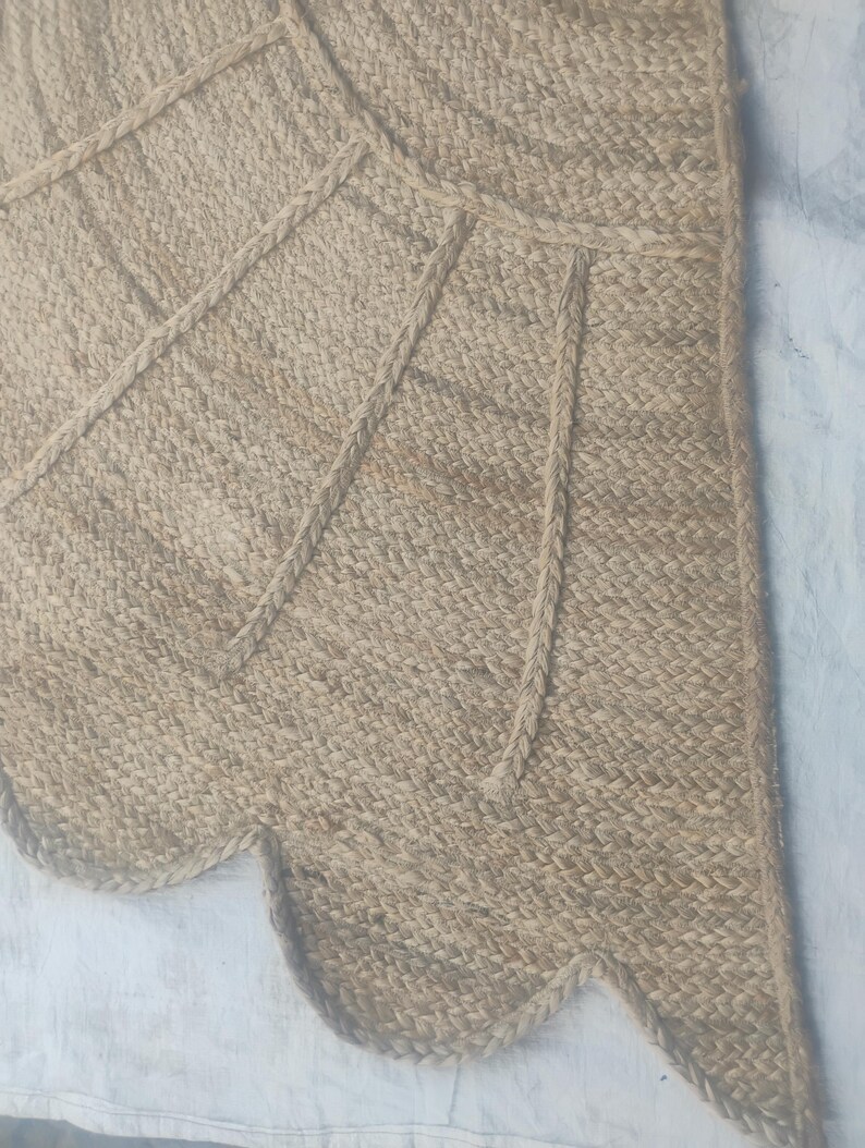 May include: A tan woven jute rug featuring a scalloped edge and a geometric pattern created by raised braided lines. The natural fibre material has a textured appearance with a light brown colour palette throughout the entire surface area.