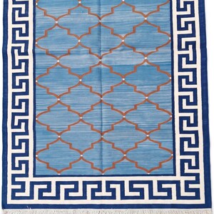 May include: A rectangular area rug featuring a light blue field with a repeating copper-coloured lattice pattern. The rug is framed by a bold navy blue and white Greek key border and finished with short white fringe along the top and bottom edges.