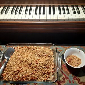 May include: A glass baking dish filled with a golden brown granola with almonds. A small white bowl with a small portion of granola is on the right side of the image.