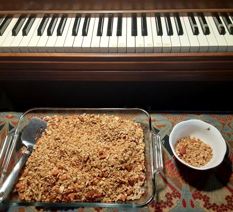 May include: A glass baking dish filled with a golden brown granola with almonds. A small white bowl with a small portion of granola is on the right side of the image.