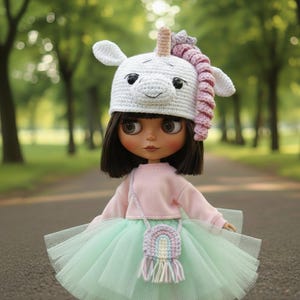May include: A doll wearing a white crochet unicorn hat with a pink and purple mane, a pink sweater, and a mint green tutu skirt. A small rainbow purse hangs from the doll's shoulder. The doll has brown hair and eyes.