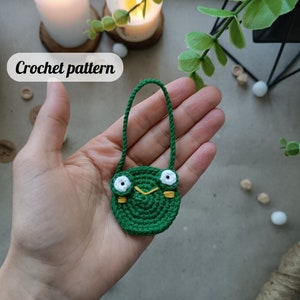 May include: A green crocheted frog-shaped purse with yellow and white details. The purse has a strap and is held in a hand.