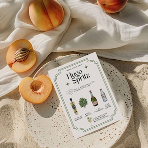 May include: A still-life image featuring a Hugo Spritz recipe card, peaches, and a plate. The recipe card lists ingredients like St. Germain, Prosecco, and soda water. Halved peaches with visible pits are arranged around the plate, with a white cloth in the background.