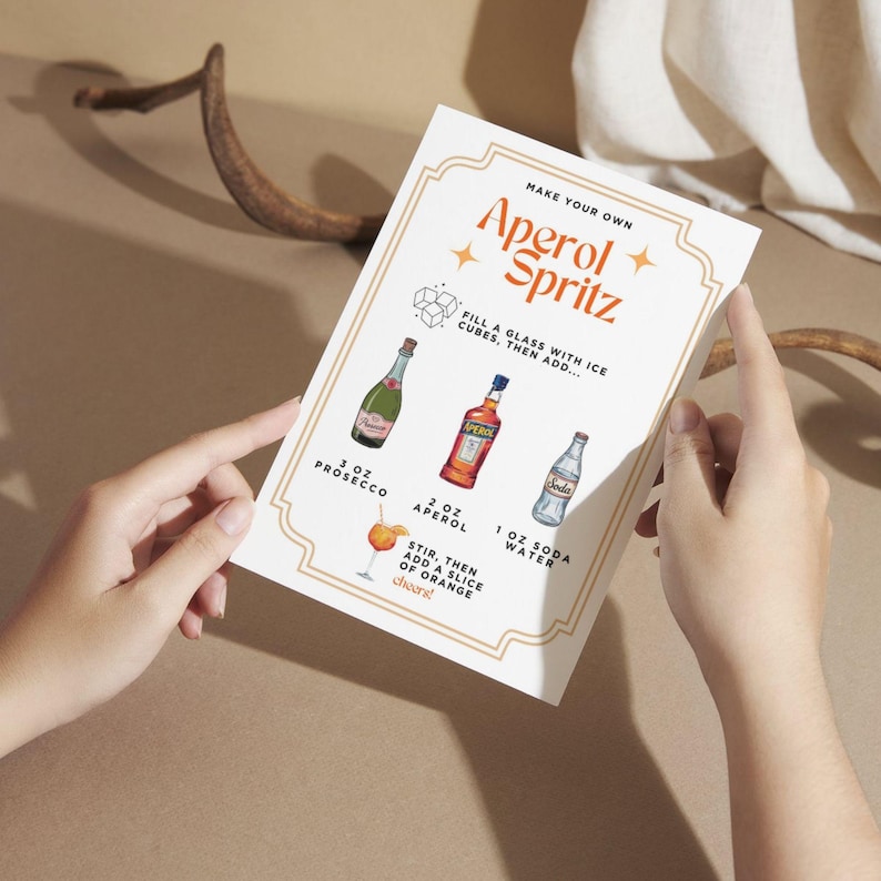 May include: A recipe card for an Aperol Spritz cocktail. The card features the title "Aperol Spritz" in a decorative font, along with instructions and illustrations of the ingredients: Prosecco, Aperol, soda water, and an orange slice.