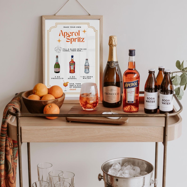 May include: Aperol Spritz cocktail ingredients arranged on a bar cart. The cart holds a framed recipe, bottles of Prosecco, Aperol, and soda water, a bowl of oranges, and a glass of the cocktail. A metal ice bucket is in the foreground.