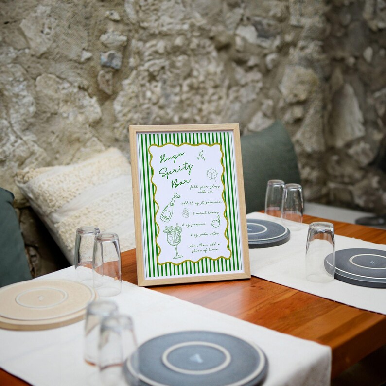 May include: A framed sign with green and white stripes that reads "Hygge Spritz Bar" with instructions for making a drink. The sign is on a wooden table with glasses and coasters.
