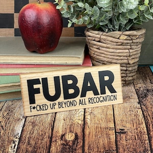 Puede incluir: Un letrero de madera con el texto "FUBAR" en letras negras y la frase "F*CKED UP BEYOND ALL RECOGNITION" en letras negras más pequeñas.