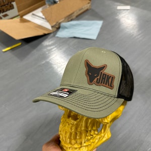 Może przedstawiać: Zielono-czarna czapka trucker z brązową skórzaną naszywką przedstawiającą czarny sylwetkę lisa i napis "JAKL".