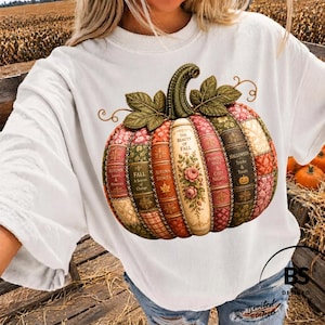 Peut inclure: Un t-shirt blanc avec un grand motif représentant une citrouille composée de dos de livres verticaux. Les livres portent des titres comme The Beauty of Fall, Autumn Stories et Halloween Tricks & Treats. La citrouille a des feuilles vertes.