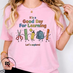 Könnte beinhalten: Ein hellrosa T-Shirt mit dem Text "it's a Good Day For Learning" und "Let's explore!" über und unter einer Grafik von animierten Schulsachen, darunter eine Schere, ein Lineal, ein Textmarker, ein Rucksack und ein Stift mit Gesichtern.