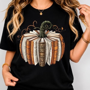 Peut inclure: Un t-shirt noir avec une impression graphique représentant une citrouille formée par des livres ouverts. Les livres ont des couvertures marron et beige avec des pages visibles, des détails de reliure et une illustration de hibou.