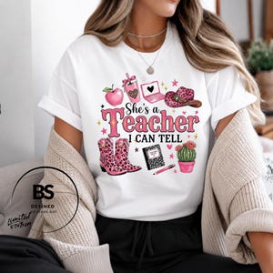 May include: A white short-sleeve t-shirt featuring a graphic print with the text "She's a Teacher I CAN TELL". The design includes a pink apple, a leopard print tumbler, a laptop, a cowboy hat, boots, a notebook, a pencil, and a potted cactus.