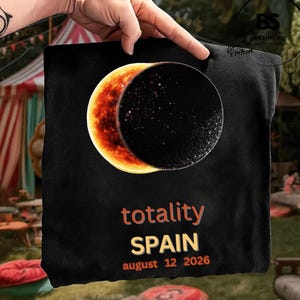 Peut inclure: Un t-shirt noir avec un graphique d'éclipse solaire montrant un croissant de soleil orange brillant et une lune sombre. Sous le graphique, le texte "totality", "SPAIN" et "august 12 2026" est imprimé en lettres orange et jaunes sur le tissu.