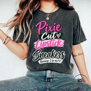 Peut inclure: Un t-shirt gris anthracite à manches courtes avec le texte Pixie Cut, Lipstick and Sneakers, Because I do me! imprimé sur le devant en lettres cursives et capitales roses, blanches et vert foncé, agrémenté de petits cœurs.