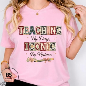 Peut inclure: Un t-shirt rose clair avec le texte "TEACHING By Day, ICONIC By Nature" écrit dans une police à blocs multicolores. Sous le texte se trouve un dessin de crayon avec un petit nœud et un bouquet de fleurs. Le t-shirt a des manches courtes.